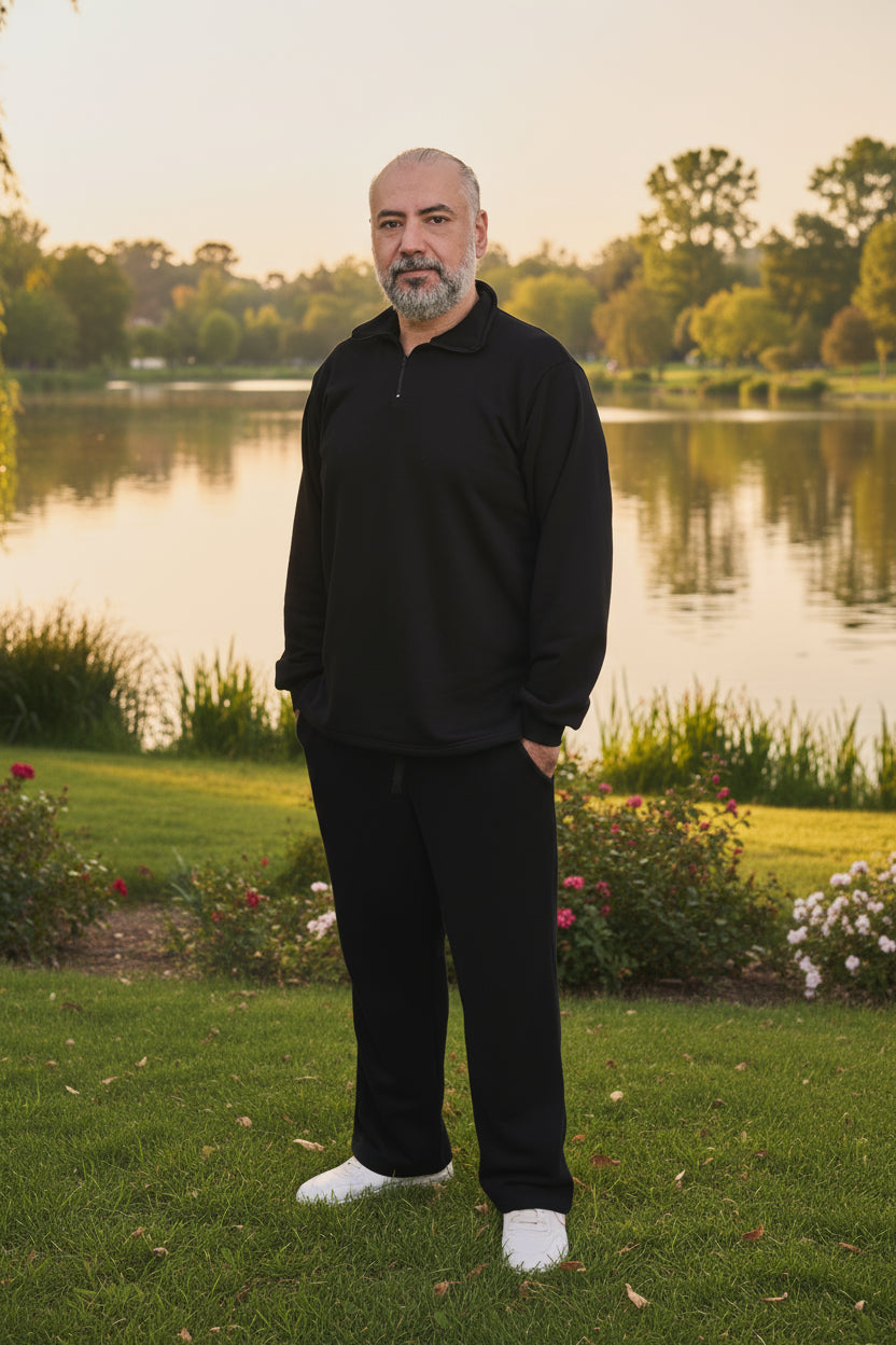 Man in black outfit standing by a lake with trees and flowers in the background